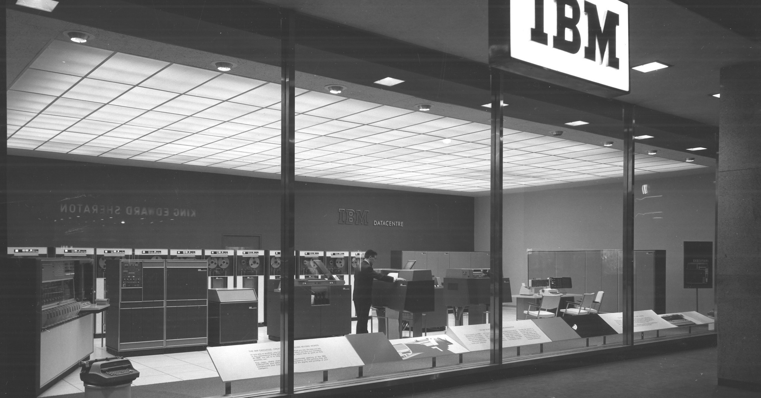 A black and white photo of a well-lit, scrupulously tidy computer room, visible through a floor-to-ceiling window. Above the window, there's a lighted IBM sign. (📸 ©International Business Machines Corporation)