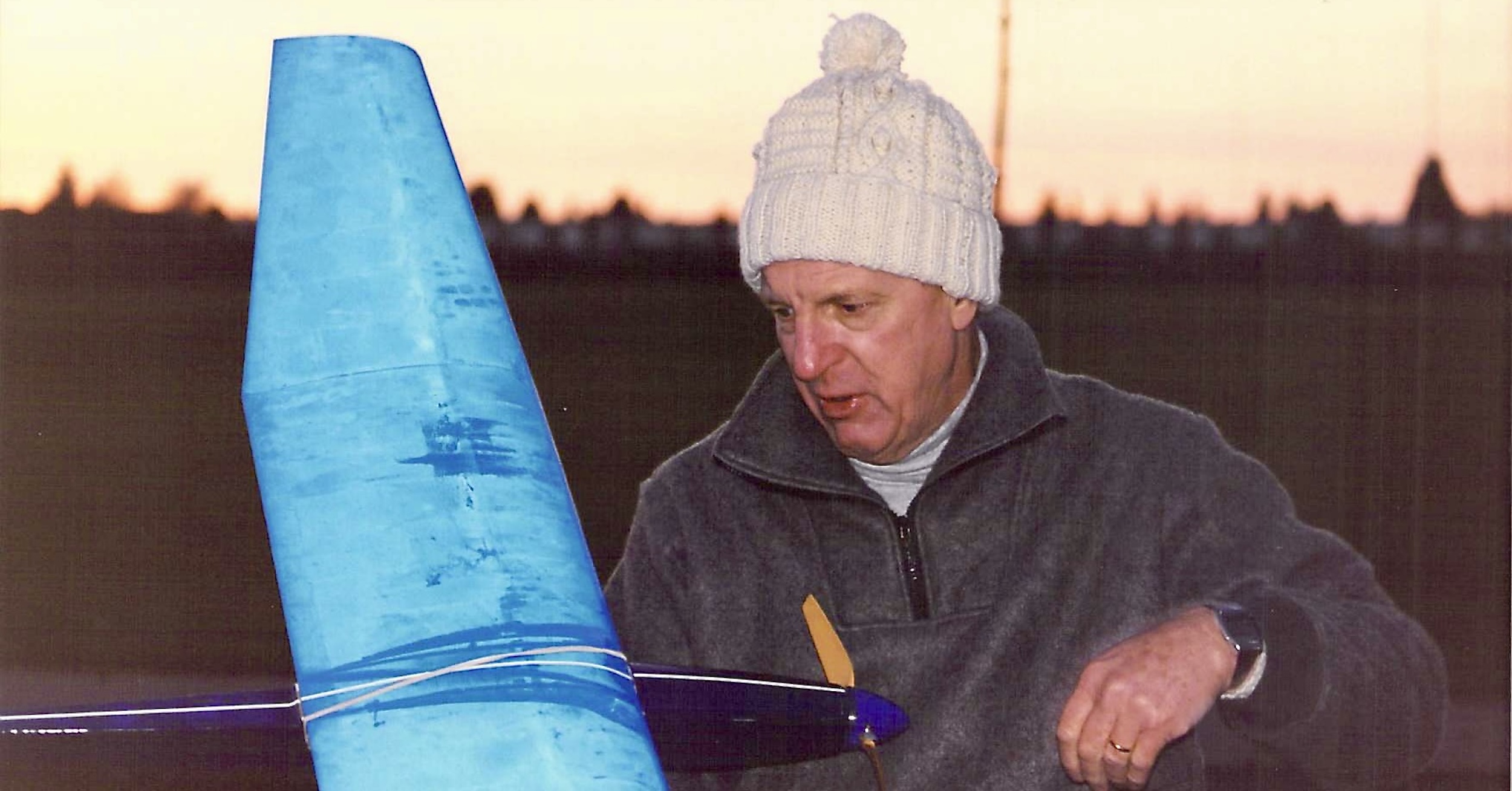 A man holds a radio-controlled model glider with a sunset sky in the background. The shiny black surface of the wing is covered in dew, which is highlighted by the camera. He is wearing a white, knitted Aran wool hat and a grey, fleece pullover. (📸 Terence C. Gannon)